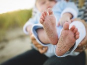 Person hält ein neugeborenes Baby auf dem Arm, der Fokus steht auf die nackten Füßchen, an denen Sand haftet. Standesamt, Anmeldung einer Geburt, Adoption, Pflegeeltern, Familie, Kindertagespflege