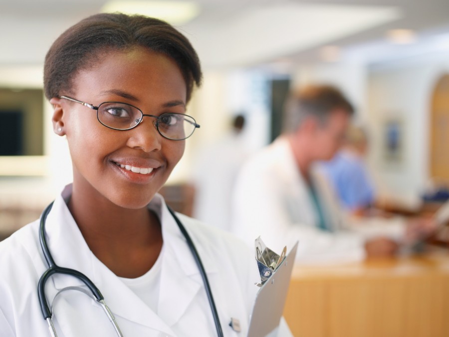 Krankenpflege, Krankenschwester, Altenpflege, Medizin: Das Fotos zeigt eine junge Frau im Arztkittel mit einem Clipboard unterm Arm in einem Krankenhaus - Canva