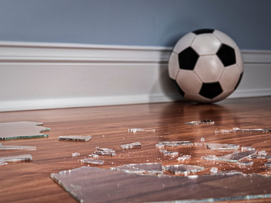 Sachschaden: Fußball und zerbrochenes Glas einer Fensterscheibe auf dem Boden in einem Raum. Infos zur Haftpflichtversicherung. - Canva